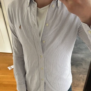 Randig skjorta från Polo Ralph Lauren - Säljer en klassisk blårandig skjorta från Polo Ralph Lauren i modellen Custom Fit. Skjortan har långa ärmar, knappar framtill och en broderad logga på bröstet. 