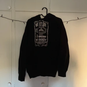 Svart hoodie med Mötley Crüe-tryck - Svart hoodie med stort vitt Mötley Crüe-tryck på bröstet inspirerat av Jack Daniel's. Klassisk huva och lång ärm, perfekt för dig som gillar rock och vill ha en bekväm tröja med attityd.(M)
