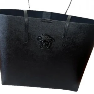 Svart handväska från Versace - Säljer en stilren svart handväska från Versace med klassisk Medusa-detalj framtill. Väskan är rymlig med två handtag och tillverkad i skinn. Perfekt för dig som vill ha en lyxig accessoar till vardagen.