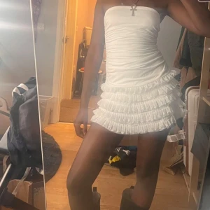 Vit axelbandslös volangklänning - Säljer en vit axelbandslös kortklänning med tight passform och flera lager volanger på kjolen. Klänningen har en rynkad överdel och är perfekt för sommarens alla tillfällen. Supersnygg och trendig modell!