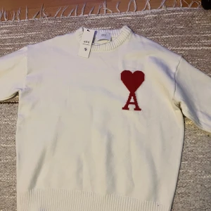 Vit stickad tröja från Ami Paris - Vit stickad tröja från Ami Paris med röd hjärt- och A-logga på bröstet. Tröjan har rund halsringning och ribbade muddar vid hals, ärmslut och nederkant. Perfekt för dig som gillar stilrena och trendiga plagg.