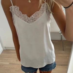 Vit linne med spetsdetaljer - Säljer ett stilrent vitt linne med vacker spetsdetalj vid halsringningen. Linnet har smala axelband och en lös passform, perfekt för varma sommardagar.💕