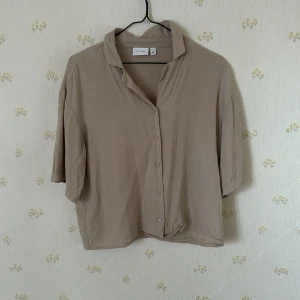  Kortärmad skjorta  - Säljer en beige skjorta från VILA med avslappnad passform och kortare ärmar. Skjortan har klassisk krage och knäppning framtill. Perfekt för en stilren och enkel look.