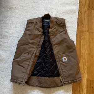 Brun quiltad väst från Carhartt - Säljer en brun väst från Carhartt med quiltat svart foder och dragkedja. Västen har två stora fickor framtill och klassisk Carhartt-logga på vänster ficka. Perfekt för lager-på-lager och stilren look.