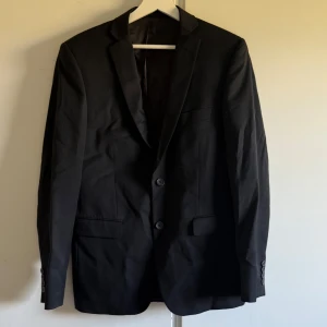 Svart kostym från Dressmann Slim Fit - Säljer en svart kostym från Dressmann i slim fit-modell, storlek 48 regular. Kavajen har två knappar framtill, klassisk krage och fickor med lock. Byxorna är raka och matchar kavajen perfekt. Perfekt för formella tillfällen. 