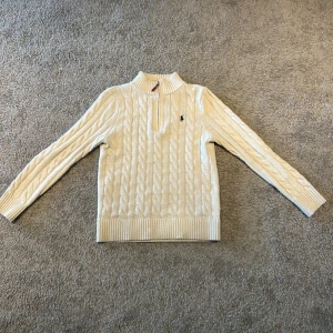 Vit kabelstickad tröja från Polo Ralph Lauren - Säljer en klassisk vit kabelstickad tröja från Polo Ralph Lauren med halv dragkedja och broderad logga på bröstet. Tröjan har lång ärm, ribbade muddar och hög krage. Perfekt för en stilren och tidlös look.