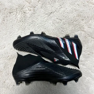 Adidas fotbollsskor i svart med vita och röda detaljer - Säljer ett par svarta Adidas fotbollsskor med vita och röda ränder på sidan. Skorna har en strumpliknande passform utan snörning och är designade för spel på gräs. Snygg och modern look med platt sula och rund tå. (Skick: 9/10)