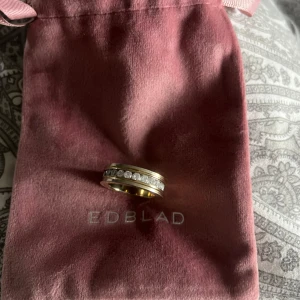 Guldfärgad ring med stenar från Edblad - Snygg guldfärgad ring från Edblad med en rad gnistrande stenar runt om. Ringen har en modern design och är perfekt för att ge din look lite extra glans. Levereras i originalpåse från Edblad. STORLEK 19