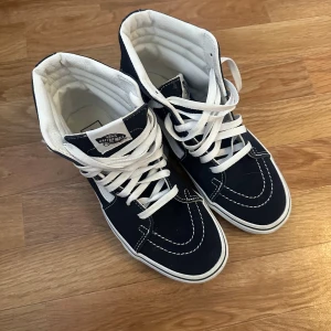 Marinblåa höga sneakers från Vans - Säljer ett par klassiska marinblåa höga sneakers från Vans med vit sula och vita snören. Skorna har den ikoniska vita Vans-sidan och är perfekta för en streetwear-look. Bekväma och stilrena skor som passar till det mesta.