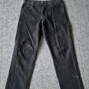 Svarta cargobyxor från Dickies - Svarta cargobyxor från Dickies med flera praktiska fickor på sidorna och bak. Byxorna har normal passform och är tillverkade i slitstark bomull. Perfekta för dig som gillar en avslappnad och funktionell stil. Storlek 32x32