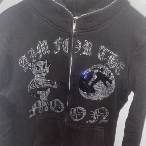Svart hoodie med strasstenar från Aim For The Moon - Svart hoodie med dragkedja och glittrande strasstenar framtill. Motiv med djävul och måne samt texten 'AIM FOR THE MOON'. Unik och edgy design, perfekt för dig som gillar statement-plagg. Alla stenar är kvar och inga synliga tecken på användning. Vid snabb affär kan pris diskuteras:)