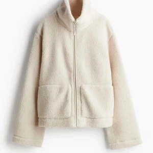 Beige teddyjacka med dragkedja - Mysig beige teddyjacka med hög krage och dragkedja framtill. Jackan har två stora fickor och är långärmad. Perfekt för kyliga dagar och ger en avslappnad look.