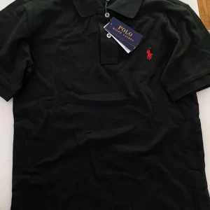 Svart pikétröja från Polo Ralph Lauren - Klassisk svart pikétröja från Polo Ralph Lauren med korta ärmar och krage. Tröjan har två knappar vid halsen och den ikoniska röda logotypen broderad på bröstet. Perfekt för en stilren och avslappnad look.