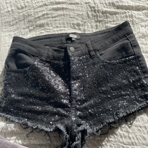 Svarta paljettshorts med fransar - Snygga svarta shorts med glittriga paljetter framtill och råa fransade kanter. Klassisk femficksmodell med knapp och dragkedja. Perfekta för att sticka ut lite extra!