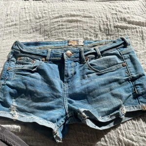 Blå jeansshorts med slitningar - Snygga blå jeansshorts med råa kanter och slitna detaljer framtill. Klassisk femficksmodell med knapp och dragkedja. Perfekta för varma dagar och enkel att matcha med olika toppar.