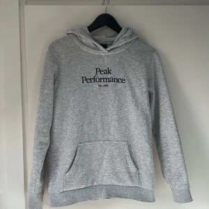 Grå hoodie från Peak Performance - Säljer en stilren grå hoodie från Peak Performance med svart broderad logga på bröstet. Tröjan har huva och en stor magficka framtill. Perfekt för en avslappnad look.