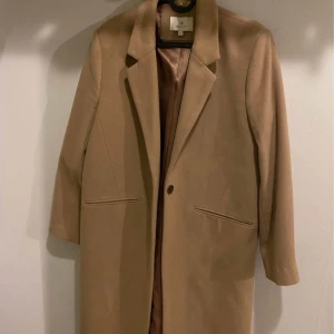 Beige kappa från & Other Stories Gant  - Stilren beige kappa med klassisk krage och en knapp framtill. Kappan har två snedställda fickor och är helfodrad. Perfekt för dig som gillar en tidlös och enkel look. 