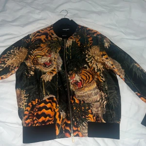 Svart bomberjacka med tigertryck från Dsquared2 - Säljer en svart bomberjacka från Dsquared2 med färgstarkt tigertryck i orange, gult och grönt. Jackan har dragkedja framtill, ribbade muddar och krage samt lång ärm. Perfekt statement-plagg för dig som vill sticka ut. 