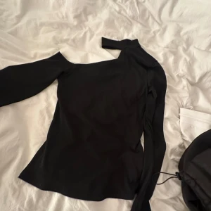 Svart oneshoulder topp med lång ärm - Säljer en svart topp med oneshoulder-design och lång ärm på ena sidan. Toppen har en snygg asymmetrisk skärning och ett band som kan knytas vid axeln för extra detalj.