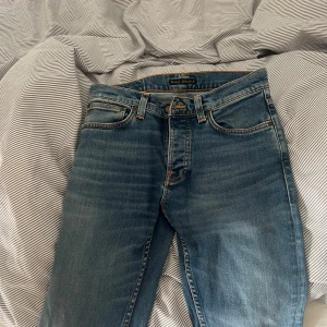 Blå jeans från Nudie Jeans - Säljer ett par klassiska blå jeans från Nudie Jeans med orangea sömmar och fem fickor. Jeansen har normal passform och stängs med knappar. Perfekta för dig som gillar stilrena och tidlösa jeans.