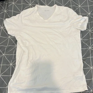 Vit t-shirt med v-ringning från Hugo Boss - Säljer en klassisk vit t-shirt från Hugo Boss med v-ringning och diskret broderad logga på bröstet. T-shirten är i mjuk bomull och har en normal passform. Perfekt basplagg till garderoben!