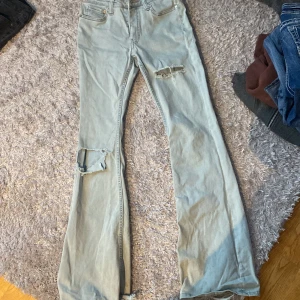 Ljusblå bootcut jeans med slitningar - Säljer ett par ljusblå jeans med bootcut-modell och slitningar på ena låret och knät. Jeansen har klassisk femficksdesign och hög midja. Perfekta för en avslappnad och trendig look.från weekday 27/34