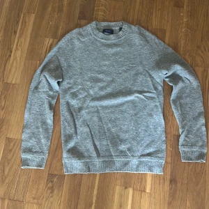 Grå stickad 100% ulltröja från GANT - Säljer en klassisk grå stickad tröja från GANT i storlek S. Tröjan har rund halsringning, ribbade muddar vid ärmslut och nederkant samt en mjuk och bekväm känsla. Perfekt för lager-på-lager under kyligare dagar.