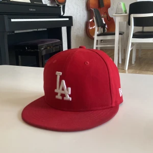 Röd LA-keps från New Era - Snygg röd keps från New Era med LA-logga i vitt framtill. Klassisk modell med platt skärm och broderade detaljer. Passar dig som gillar sportig stil och vill sticka ut med en färgstark accessoar.