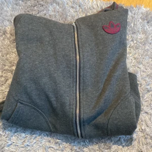 Grå hoodiejacka från Adidas - Grå hoodiejacka med dragkedja från Adidas. Jackan har lila Adidas-logga på bröstet och lila ränder längs ärmarna. Mjuk bomullskänsla och fickor framtill. Perfekt för en sportig och avslappnad stil. 