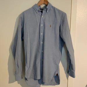 Ljusblå skjorta från Ralph Lauren - Klassisk ljusblå skjorta från Ralph Lauren med button-down krage och broderad logga på bröstet. Skjortan har lång ärm och vita knappar framtill. Perfekt för en clean och stilren look.