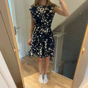 Mörkblå klänning med fjärilsmönster - Supersöt mörkblå kortklänning med vita fjärilar, markerad midja och knytband bak. Klänningen har dragkedja i ryggen, rund halsringning fram och v-ringad rygg. Är i storlek M, men passar även S finns knyte i midjan. Jag har storlek S på bilden. 