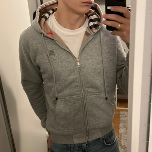 Grå Burberry zip hoodie - Snygg grå Burberry zip hoodie som har blivit lite liten för mig. Storleken är L men tycker själv att den är liten i passformen. Den är i mycket bra skick då jag har använt den med försiktighet. Har ni några frågor så är det bara att skriva. Priset går även att diskutera!