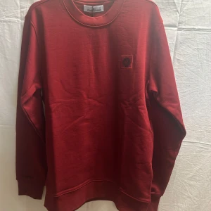 Röd sweatshirt från Stone Island.  - Snygg röd sweatshirt från Stone Island med klassisk rund hals och broderad logotyp på bröstet. Tröjan har långa ärmar och ribbade muddar vid ärmslut och nederkant.                   Obs! Skriv önskad storlek efter lagd beställning. Alla storlekar finns. 