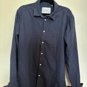 Mörkblå skjorta från Les Deux - Säljer en stilren mörkblå skjorta från Les Deux i storlek M. Skjortan har klassisk krage, vita knappar framtill och långa ärmar. Perfekt för dig som vill ha något stilrent till sommaren.