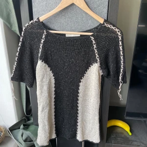 stickad topp secondhand - Säljer en unik stickad topp secondhand i svart med vita paneler och dekorativa sömmar längs axlar och sidor. Modellen har korta ärmar och rund halsringning. 