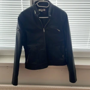 Svart skinnjacka från IsoLina - Snygg svart skinnjacka från IsoLina i storlek 40. Jackan har flera dragkedjor framtill, inklusive sned bröstficka och två sidofickor. Klassisk krage och långärmad modell, perfekt för en cool och stilren look.