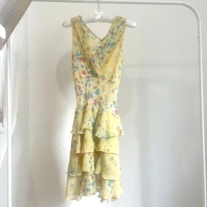Gul blommig volangklänning - Söt gul klänning med blommigt mönster i gul. Klänningen har v-ringning, är ärmlös och har flera volanger nedtill för en lekfull look. Ryggen är lite  genomskinlig (se bild 4), jämfört med brösten (bild 3).