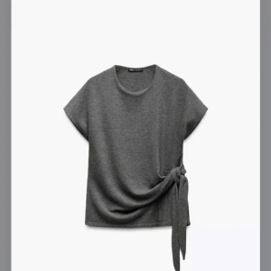Grå topp med knyt från Zara - Snygg grå topp från Zara med korta ärmar och en stilren knytning på sidan. Perfekt för en chic och avslappnad look. Tillverkad i ett mjukt material som ger en bekväm passform.