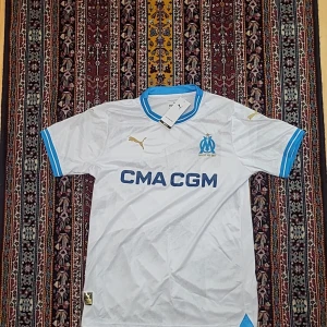 Marseille Puma vit matchtröja M - Olympique de Marseille hemmatröja från Puma i vitt med blå detaljer på krage och ärmslut. Guldigt Puma-logo på bröstet, klubbmärke och sponsortryck i blått. Tillverkad i lätt och ventilerande polyester, perfekt för fotboll eller supporterliv.