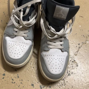 Nike Air Jordan 1 i grått och vitt - Säljer ett par Nike Air Jordan 1 i grått och vitt. Skorna har klassisk design med perforerad tå, vita snören och hög komfort. Perfekta för dig som gillar streetwear och sneakers med ikonisk stil.
