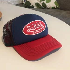 Blå och röd keps från Von Dutch - Säljer en snygg truckerkeps från Von Dutch i blått och rött med broderad logga framtill och mesh på sidorna. Justerbar baktill för perfekt passform. Kepsen har även en liten Von Dutch 99-detalj på sidan.