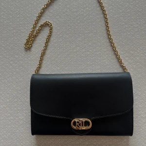 Svart kuvertväska från Ralph Lauren - Snygg svart kuvertväska från Ralph Lauren med guldfärgad kedja och RL-logga framtill. Väskan är i skinn och har flera fack inuti samt dragkedja. Perfekt för att bära det viktigaste med stil.(väskan är aldrig använd, så den är i ett väldigt fint skick🥰). Skrev till mig o du har frågor eller vill ha fler bilder.