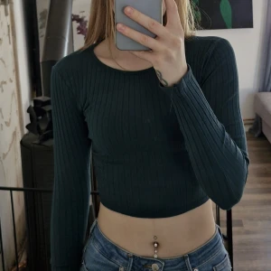 Mörkgrön ribbad croppad topp med knyt i ryggen - Trendig mörkgrön croppad topp från New Yorker med lång ärm och ribbad struktur. Toppen har snygg öppen rygg med knytband som ger en unik detalj. Perfekt för dig som gillar stilrena men ändå speciella plagg.