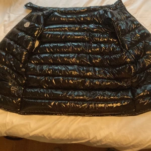Svart dunväst från Moncler - Säljer en glansig svart dunväst från Moncler med klassisk logga på bröstet. Västen har dragkedja framtill och två fickor med dragkedja. Perfekt för lager-på-lager och riktigt snygg till streetwear-stilen.
