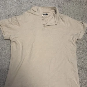 Beige pikétröja från 157, storlek S - Säljer en stilren beige pikétröja från 157 i storlek S. Tröjan har klassisk krage, korta ärmar och knappar framtill. Perfekt basic-plagg i mjuk bomull som funkar till många olika outfits.