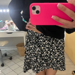 Svart blommig kjol med volang - Säljer en svart omlottkjol med vitt och gult blommönster. Kjolen har volangdetalj längs nederkanten och knyts i sidan. Perfekt för dig som gillar en feminin och trendig stil.