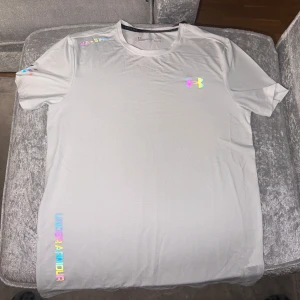 Vit t-shirt från Under Armour - Säljer en vit t-shirt från Under Armour med färgglada loggor och textdetaljer. T-shirten har rund halsringning och korta ärmar. Perfekt för träning eller sportiga outfits. Finns även i svart