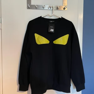 Svart Fendi tröja med gula ögon - Cool svart tröja från Fendi med ikoniska gula ögon framtill. Tröjan har rund hals, långa ärmar och en relaxed passform. Perfekt för dig som vill sticka ut med en statement piece från ett lyxigt märke. Pris kan diskuteras vid snabb affär! Tar emot byten👌