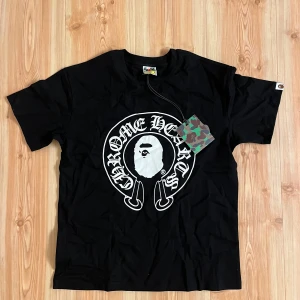 Svart BAPE x Chrome Hearts t-shirt - Svart t-shirt från BAPE x Chrome Hearts med stor vit logga och ikoniskt ape-motiv på framsidan. Baksidan har Baby Milo-tryck och Chrome Hearts-kors i vitt. Klassisk passform, kortärmad och i mjuk bomull. Perfekt för dig som gillar streetwear och unika samarbeten.