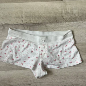 Vita pyjamas shorts med rosa rosett-mönster - Söta vita shorts med små rosa rosetter och knappar framtill. De har låg midja och är gjorda i mjuk bomull, perfekta för chill dagar hemma eller som pyjamasshorts. Stretchig midja för extra komfort och en feminin vibe. Från ginatricot och den är helt slutsåld